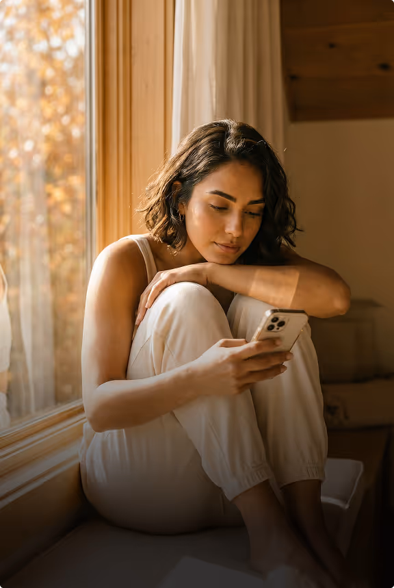 Young woman with short curly hair sitting by a window, looking thoughtfully at her smartphone.