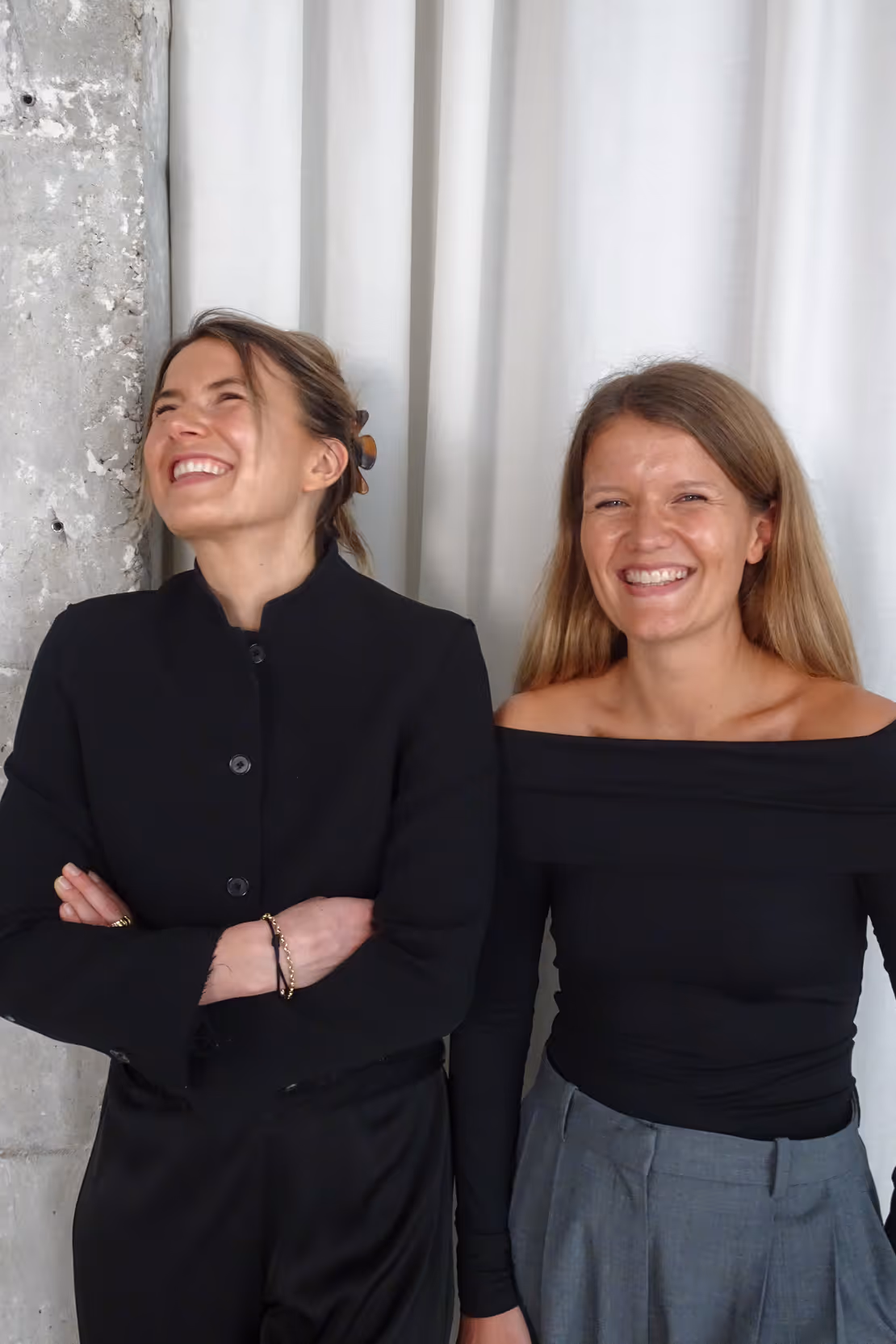 Two women smiling and standing side by side against a white curtain and concrete wall background, both wearing black tops.