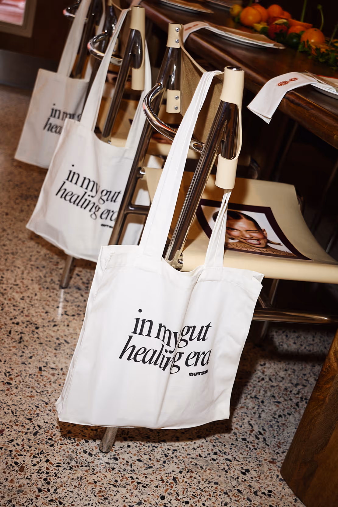 White tote bags hanging on the back of beige chairs, each bag printed with the phrase 'in my gut healing era.'