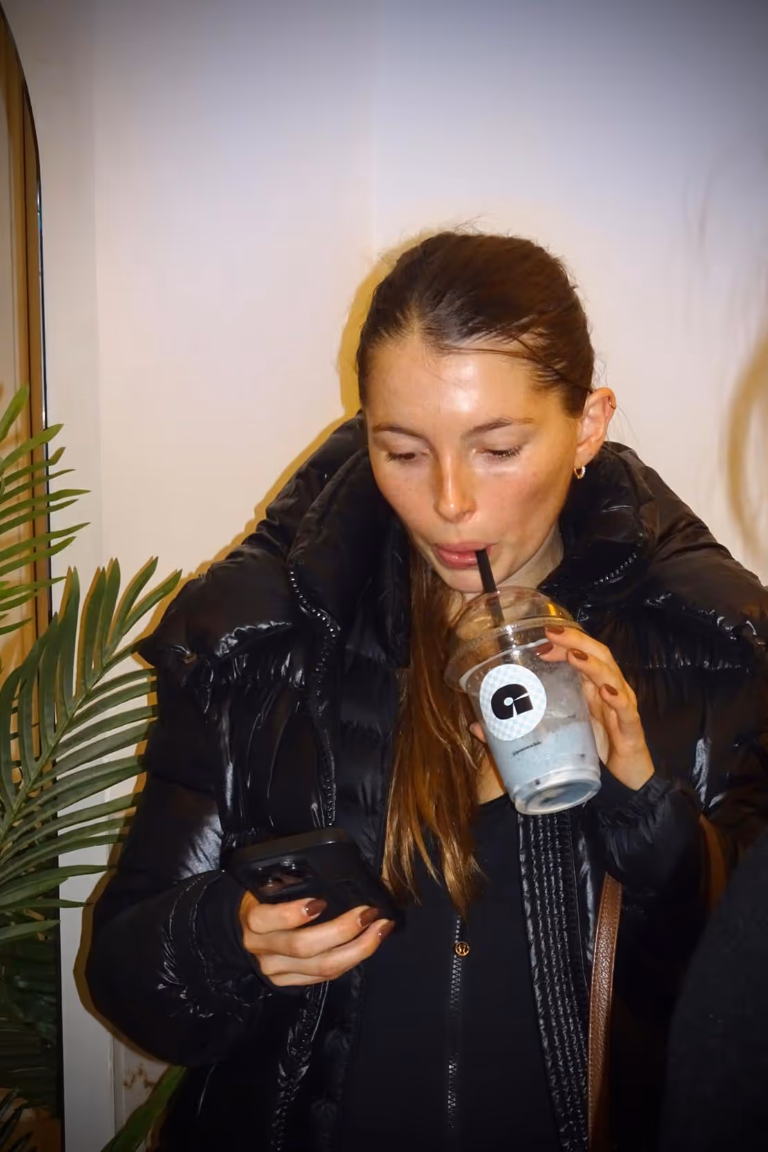 Woman in a black puffer jacket drinking from a plastic cup with a straw while looking at her phone.