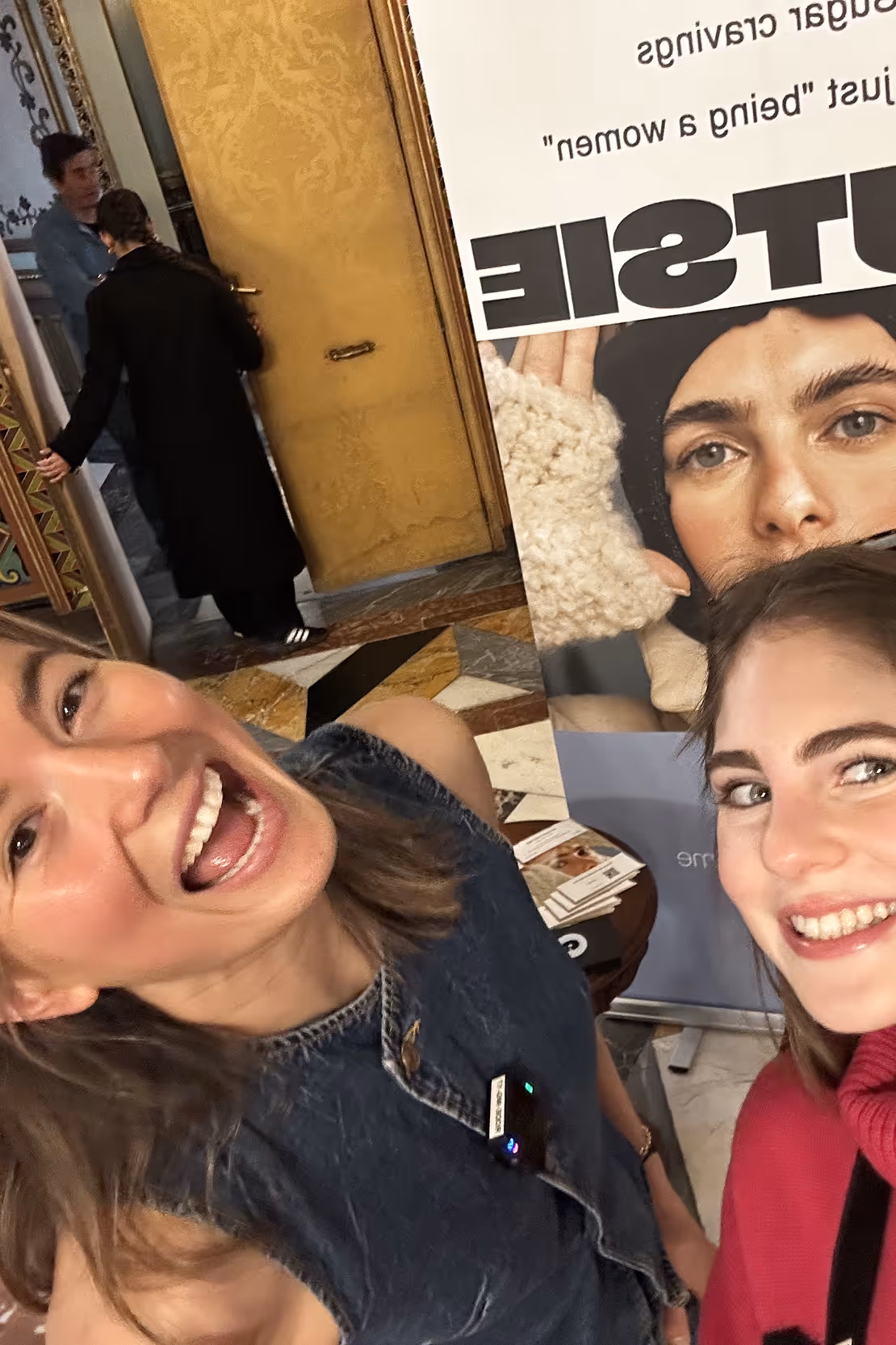 Two smiling women taking a selfie indoors in front of a poster featuring a woman's face and text.