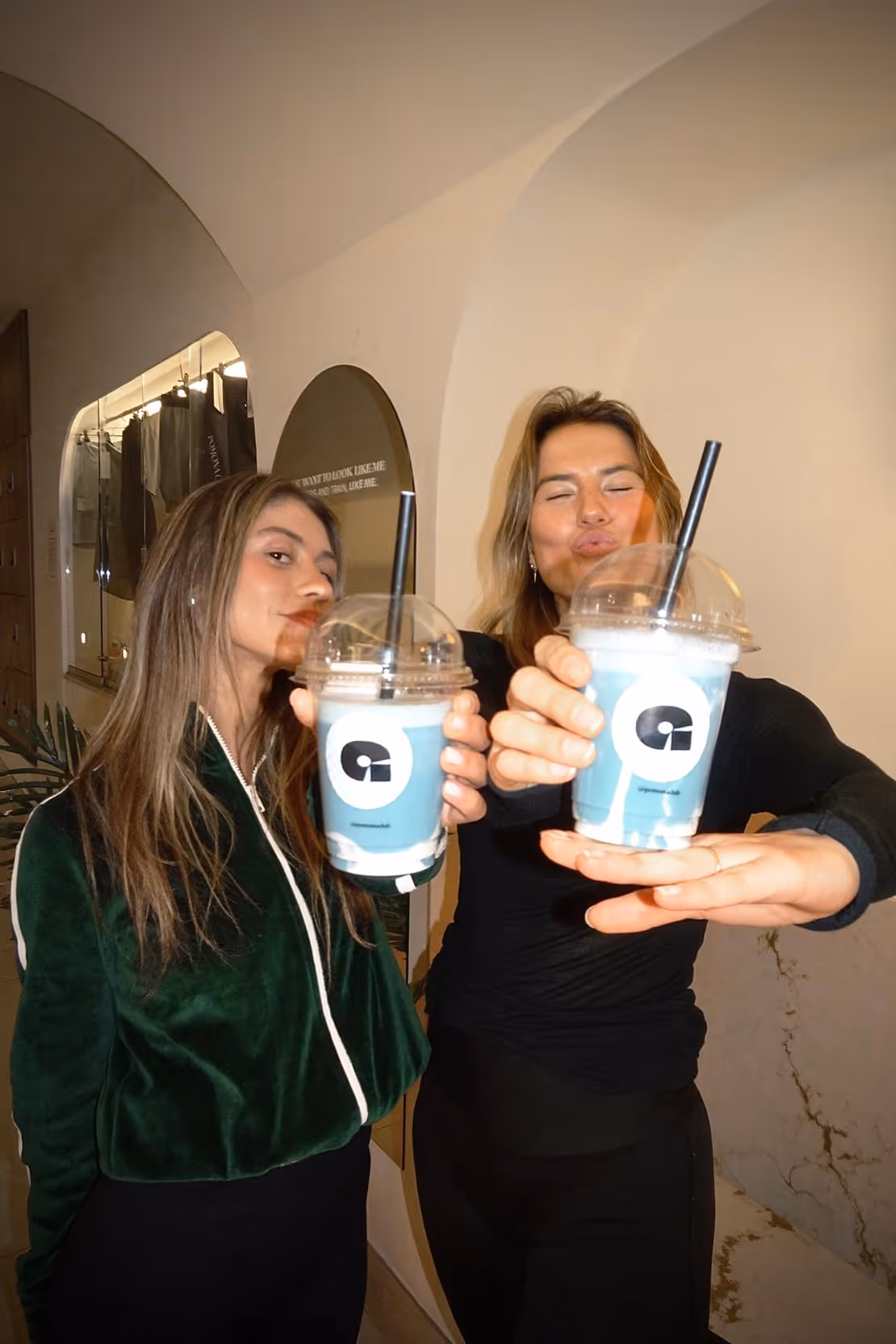 Two women holding up plastic cups with blue and white drinks and black straws, one woman is puckering her lips.