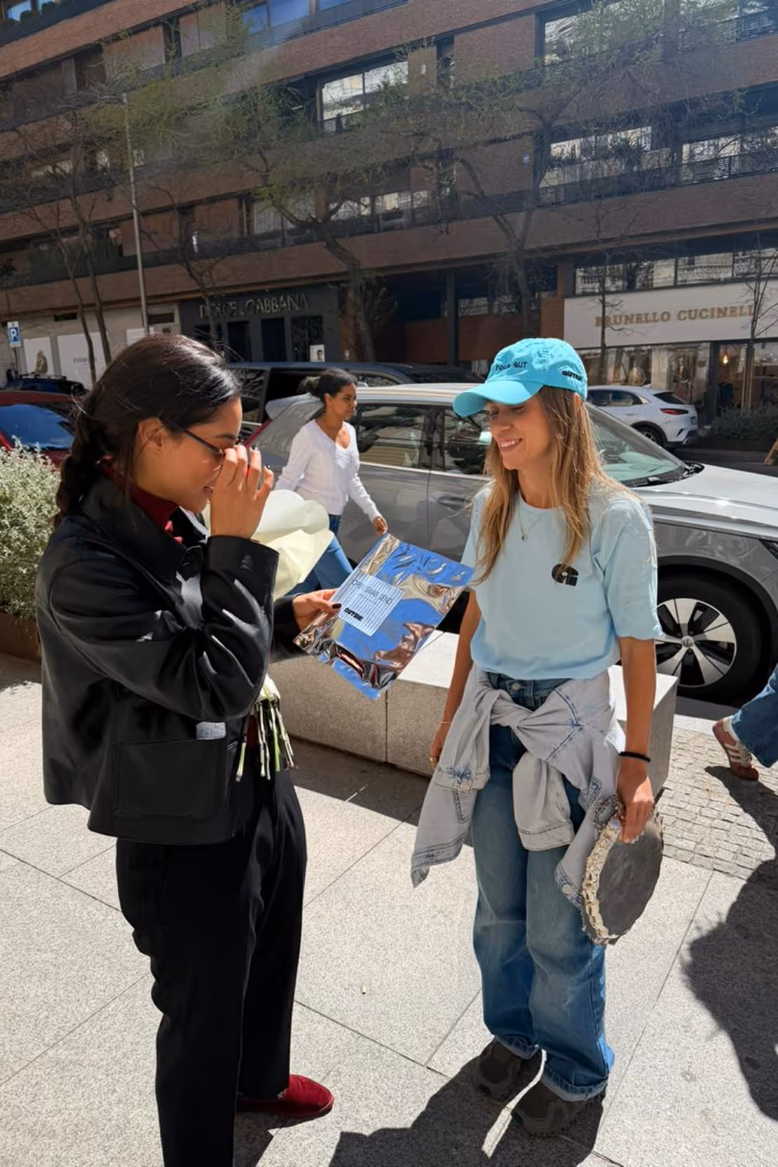 Two women standing on a sunny sidewalk, one holding flowers and a silver package, the other wearing a blue cap and holding a tambourine.