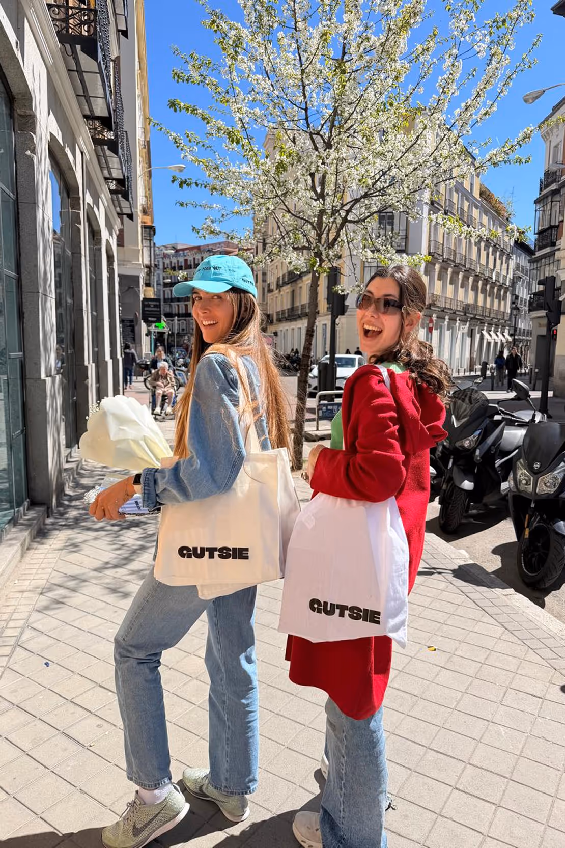 Two smiling women standing on a sunny urban sidewalk, each holding a white bag with the word GUTSIE, one wearing a blue cap and denim outfit, the other in a red coat and sunglasses.