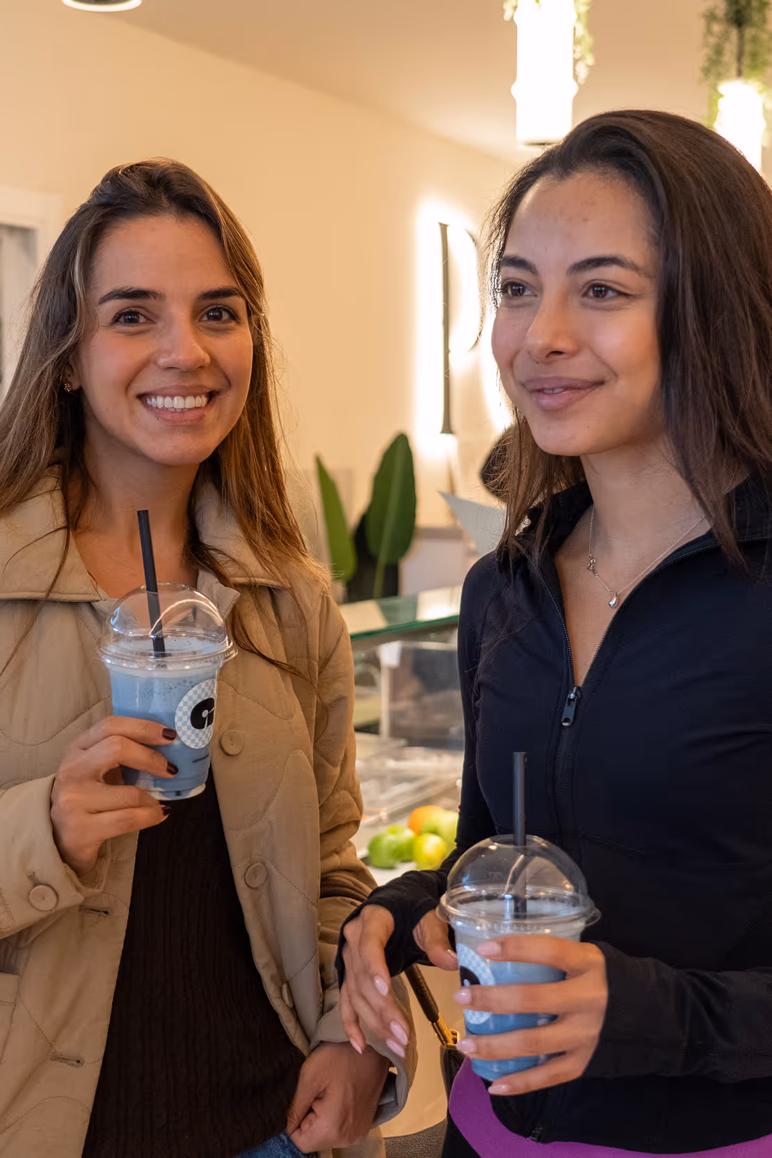 Two women smiling and holding plastic cups with blue bubble tea inside, standing indoors.