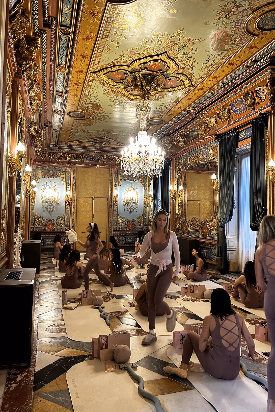 Group of women in matching brown workout outfits on yoga mats preparing for a session in an ornate room with painted ceiling and chandelier.