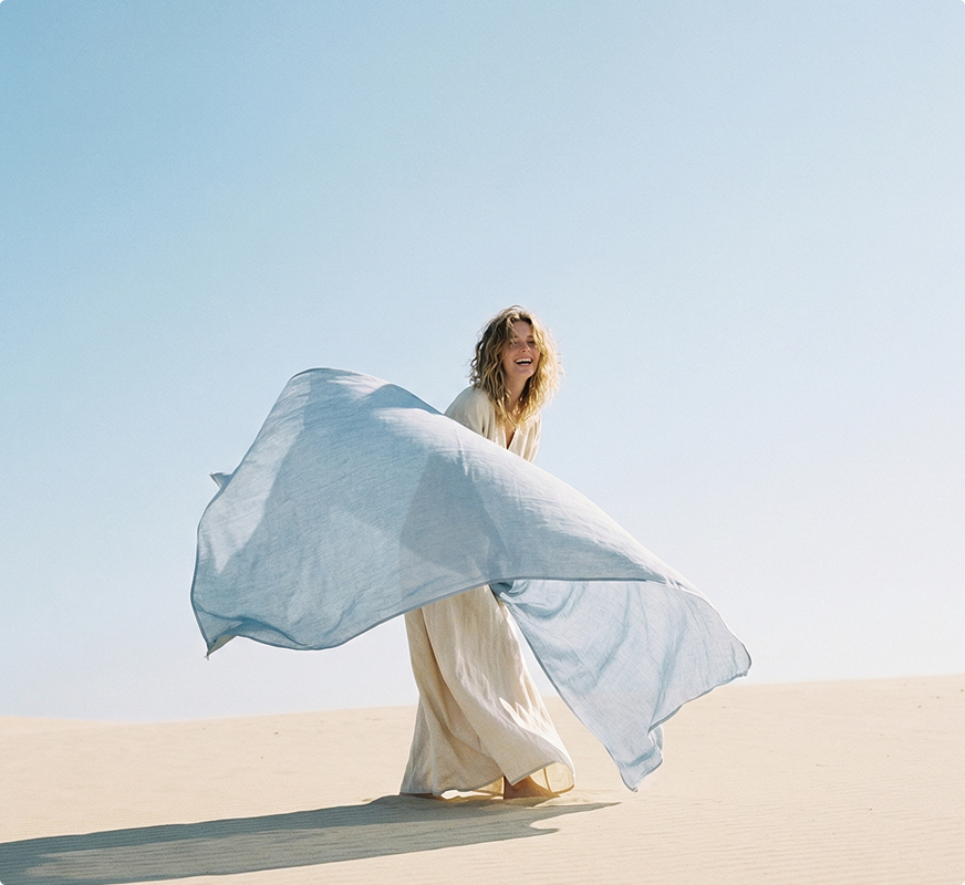 Woman in a long beige dress joyfully holding and waving a large light blue scarf in a sunny desert landscape.