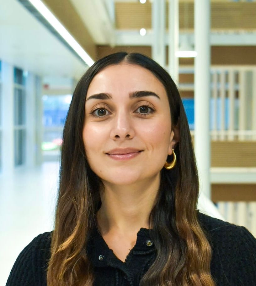 Smiling woman with long brown hair wearing a black top and gold hoop earrings in a modern indoor setting.