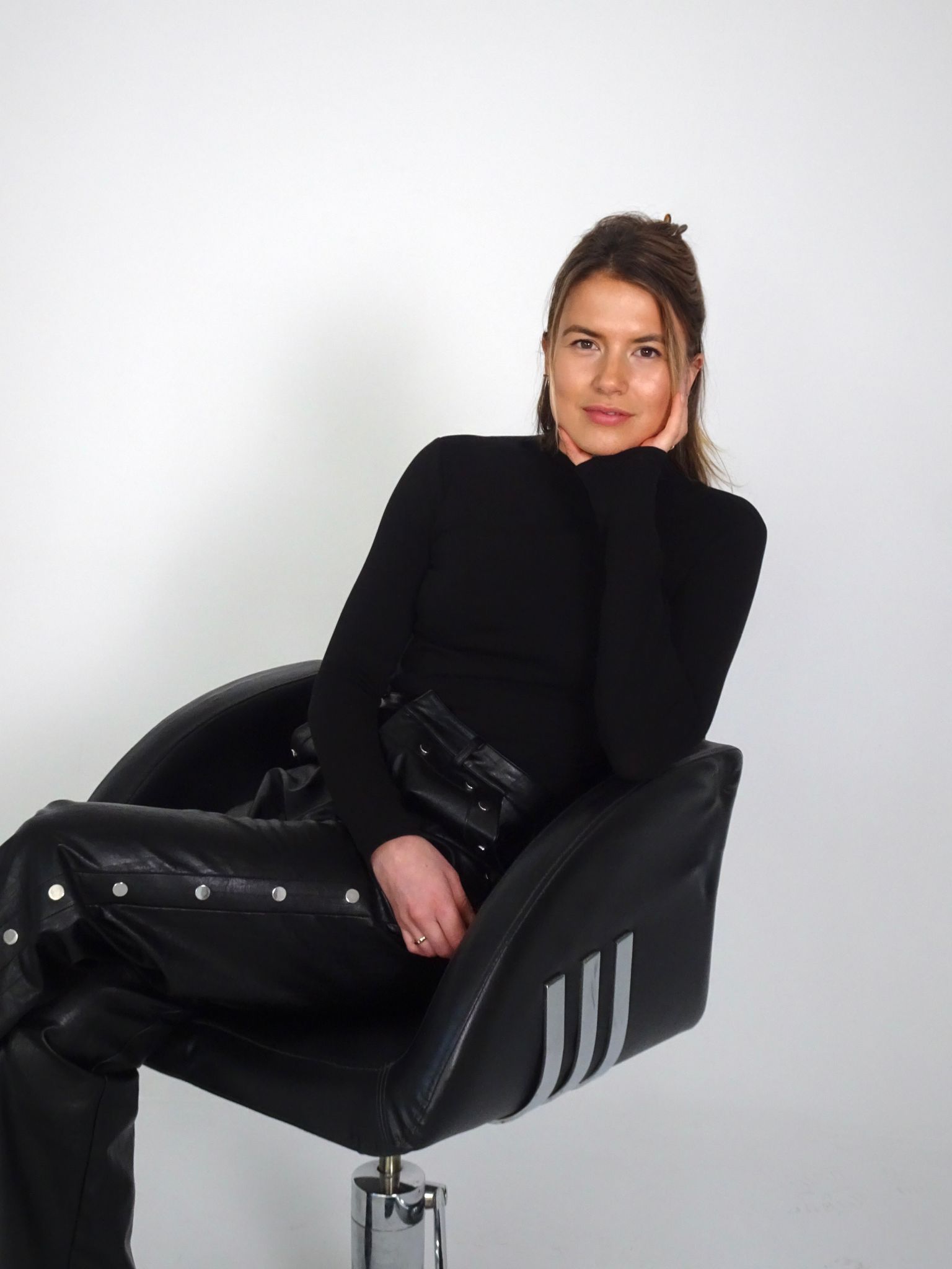 Woman with light brown hair tied back, wearing a black long-sleeve top and black leather pants with silver buttons, sitting on a black swivel chair against a plain white background.