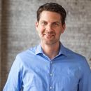 Smiling man with dark hair wearing a blue button-up shirt in front of a gray brick wall.