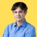 Man with dark hair wearing a blue button-up shirt against a yellow background.