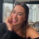 Smiling young woman with brown hair sitting indoors near a window, resting her chin on her hand.