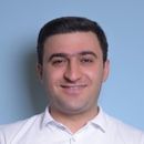 Smiling man with dark hair wearing a white collared shirt against a light blue background.