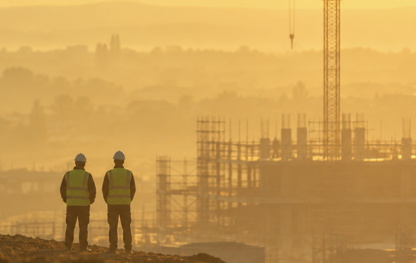 Two workers in high-visibility vests and hard hats overlooking a construction site at sunrise