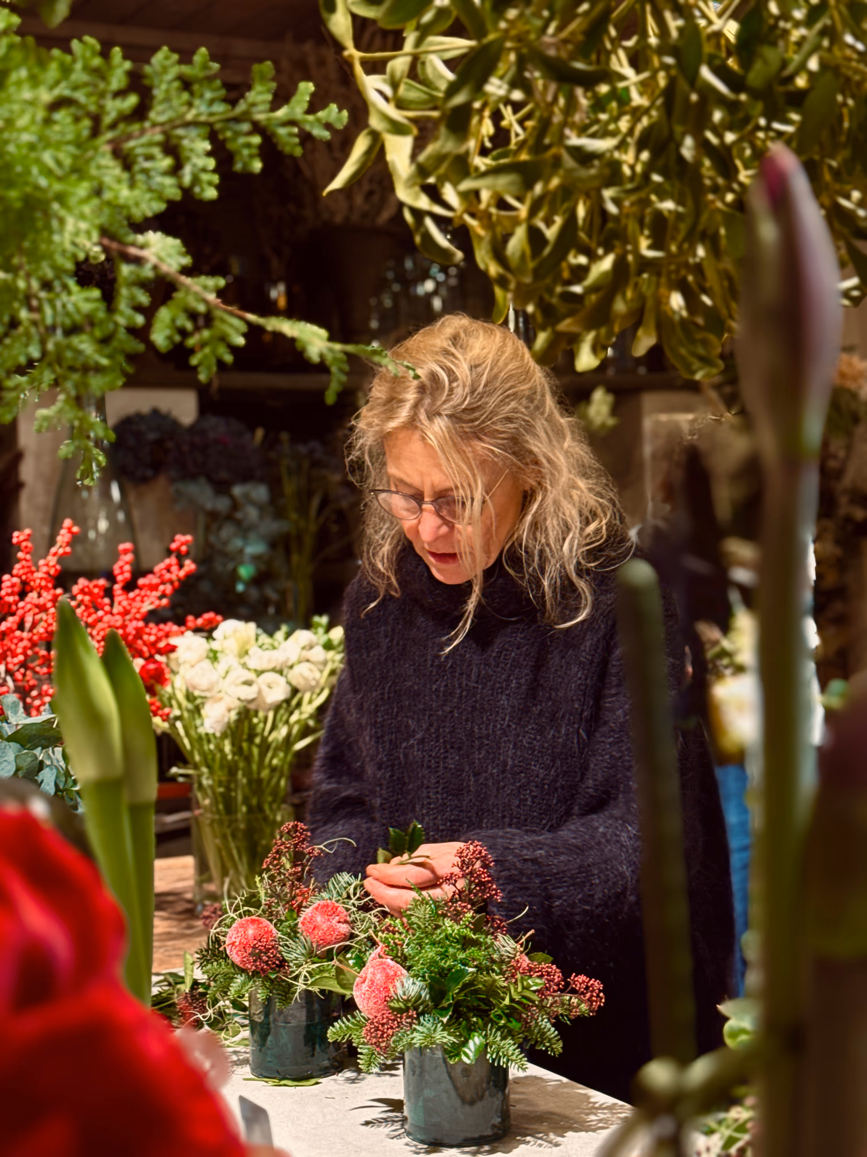 Kvinna arrangerar blommor i två glasvaser på ett bord omgivet av olika växter och blommor.