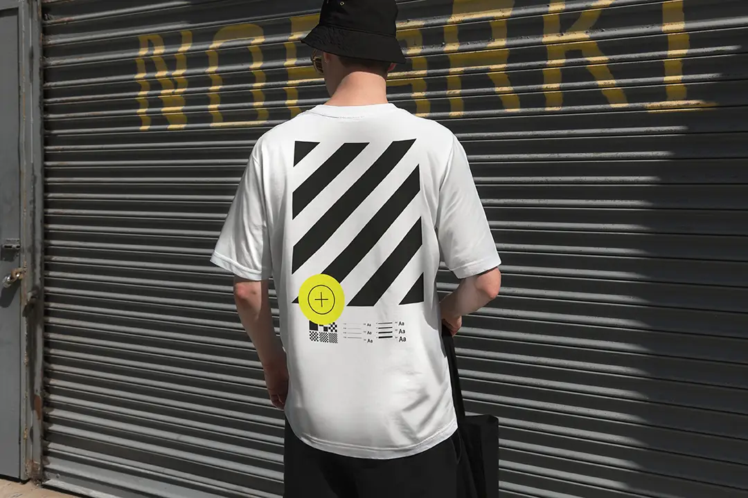 Showcase your designs with this urban streetwear mockup featuring a model standing in front of an industrial garage door. Customize the t-shirt, tote bag, and hat with any color or design to bring your branding to life. Perfect for apparel and accessory brands.