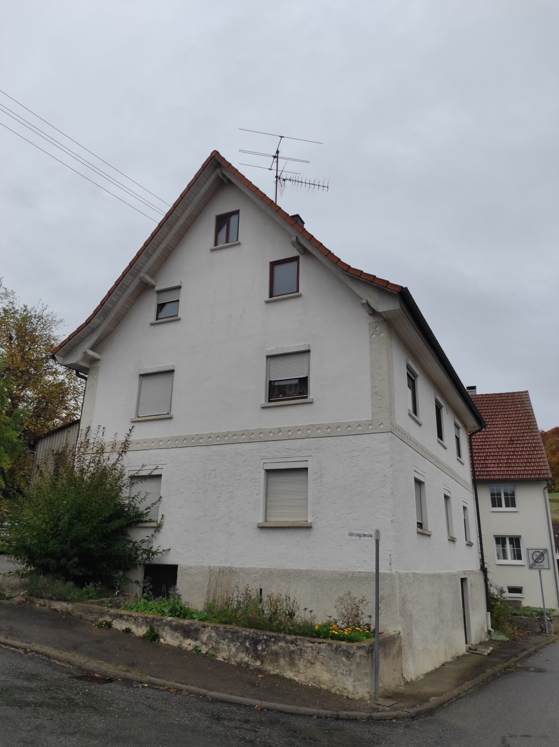 Weißes Wohnhaus mit steilem Satteldach an einer Straßenecke unter grauem, bewölktem Himmel.