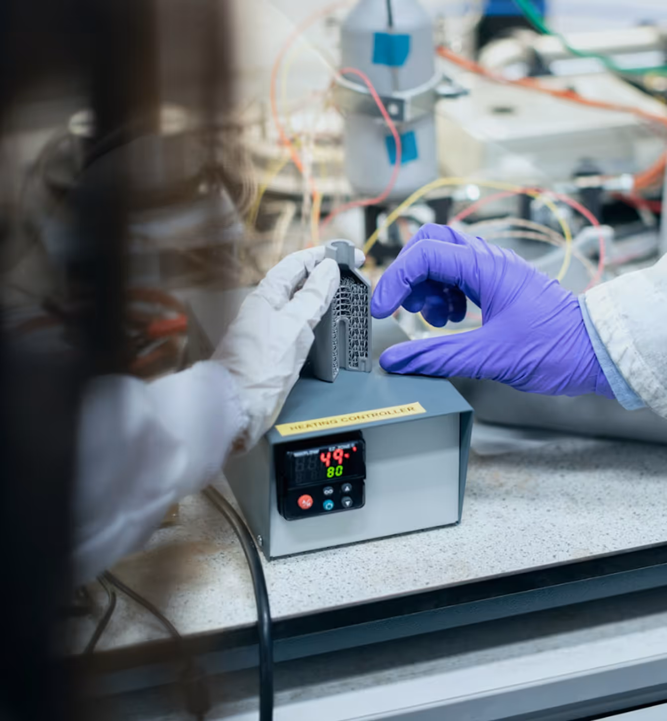 Hands wearing lab gloves adjusting a metal part on a heating controller device in a laboratory setting.