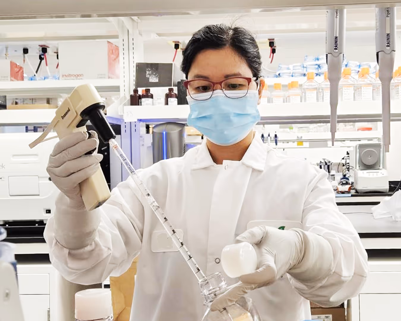 An asian woman in scrubs, wearing laboratory scrubs, face mask and red glasses, standing in laboratory. Her hands have medical gloves on them and she is holding laboratory equipment in her right hand that is pouring liquid into transparent glass bottle.