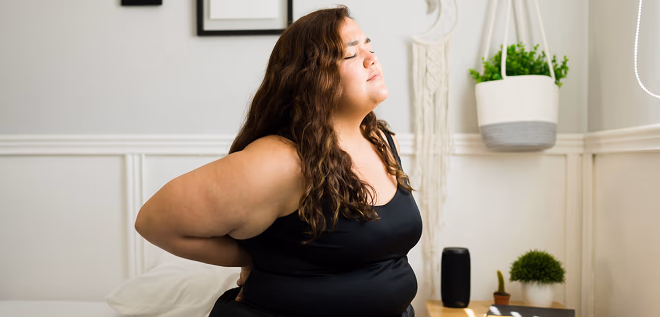 Woman in black tank top sitting on bed with eyes closed, holding her lower back in discomfort.