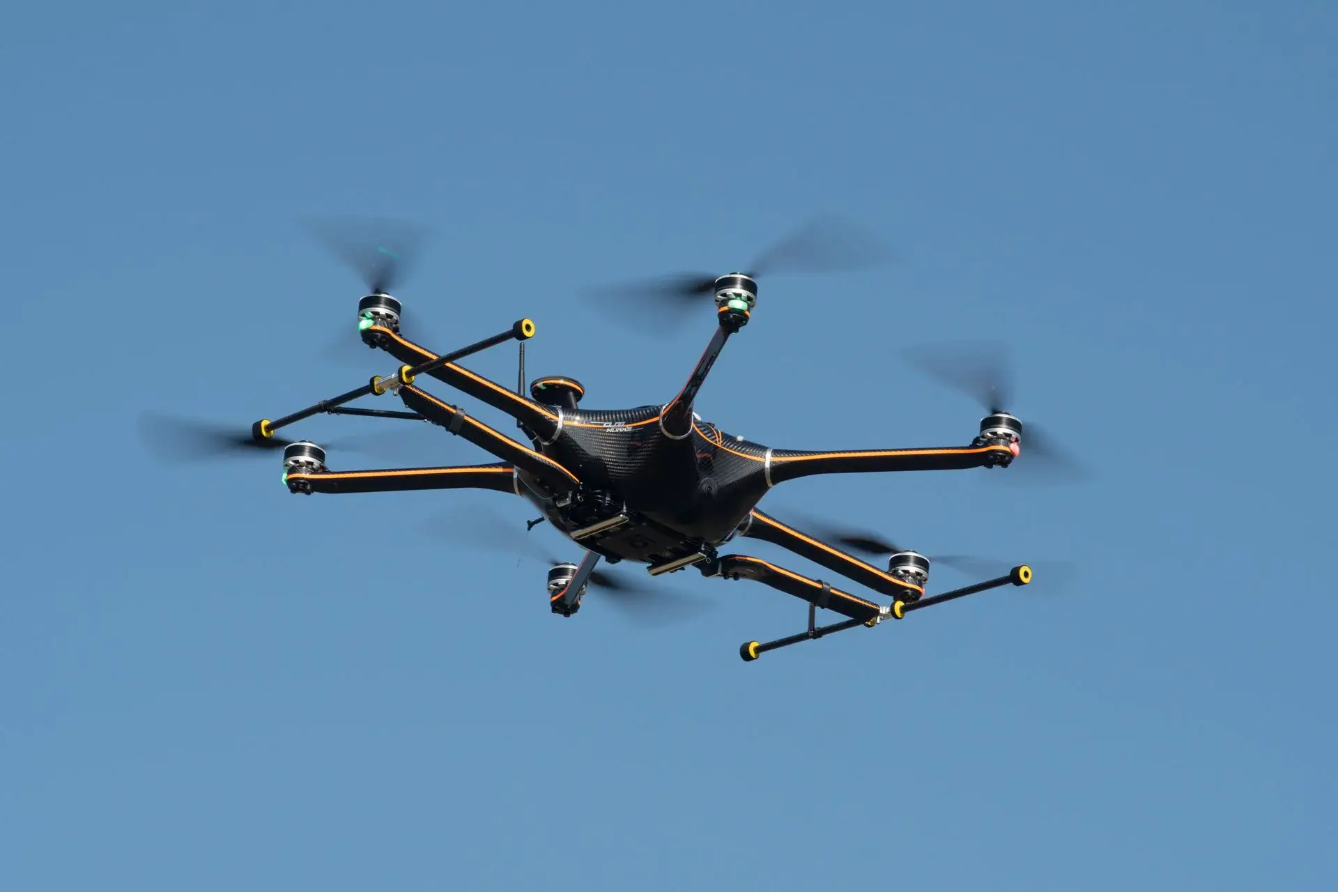 Black and orange octocopter drone flying against a clear blue sky.