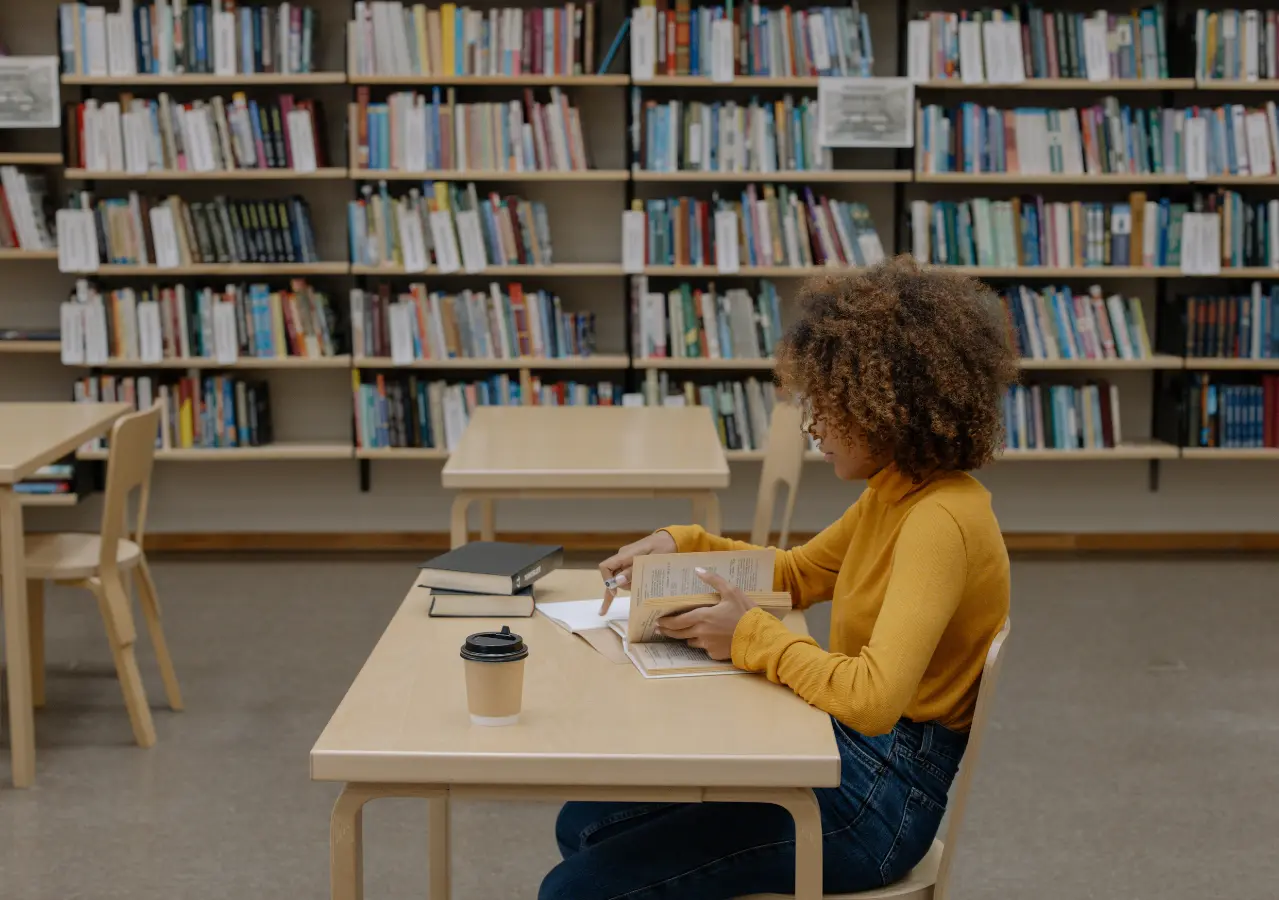 Person in a mustard yellow sweater reading a book at a wooden table in a library with shelves of books behind.