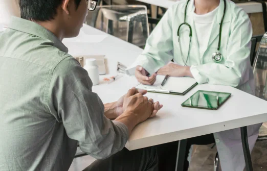 Patient sitting across from a doctor with a stethoscope at a white table during consultation.