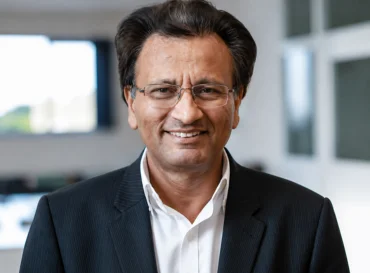 Professor Humayun Mughal with glasses and dark hair wearing a black suit jacket and white shirt, smiling indoors.