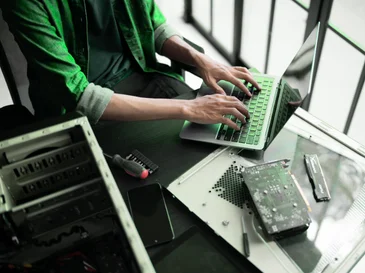 Person typing on a laptop near computer parts and tools on a desk by a window.