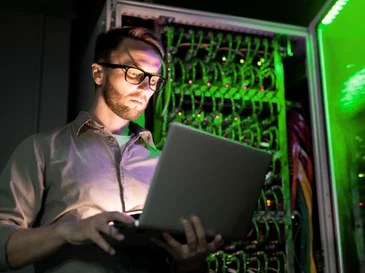 Man in glasses working on a laptop near a server rack with green lights in a dim room.