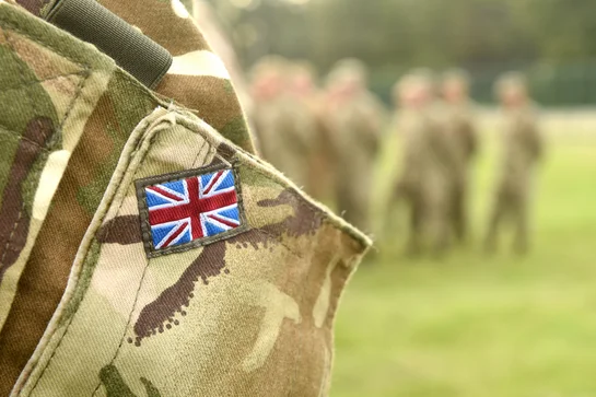 Close-up of British flag patch on camouflage uniform with blurry soldiers in background on green field.