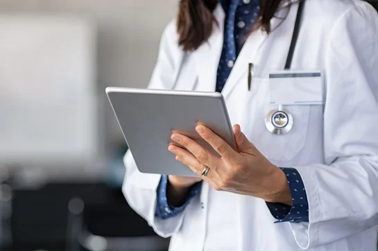 Healthcare professional in white coat using a tablet, with stethoscope around the neck.