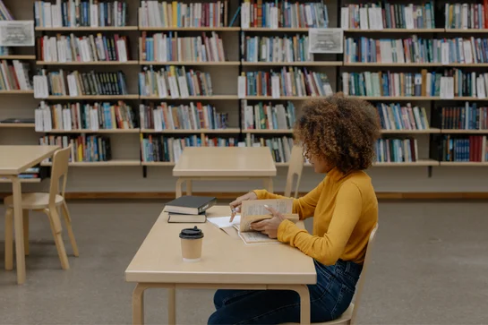 Person in a yellow sweater reading a book at a table in a library with bookshelves in the background.
