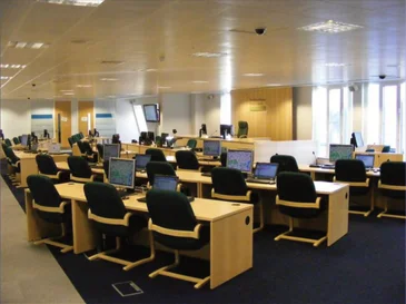 Office room with rows of desks and black chairs, each desk equipped with a computer monitor.