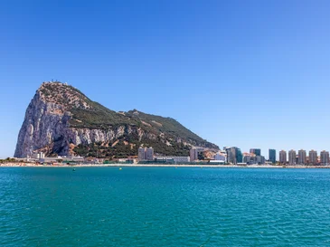 View of Gibraltar's rocky limestone promontory with city buildings along the coast under clear blue sky.