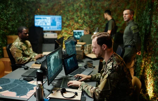 Military personnel analyzing digital maps and data on computers in a camouflaged operations room.