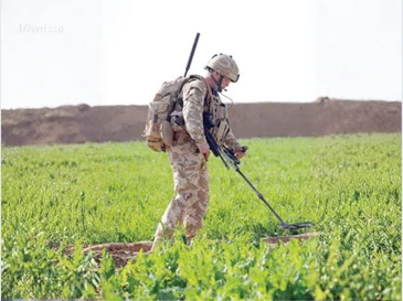 Soldier in camouflage using a metal detector to scan soil in a green field.
