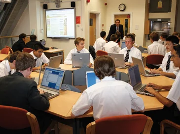 Students in school uniforms using laptops around tables while a teacher stands near a projector screen.