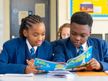 Two students in blue uniforms reading a colorful book together at a classroom desk.