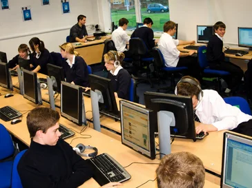 Students working on desktop computers in a classroom with large windows and multiple rows of desks.