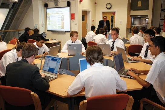 Students in white shirts seated around tables using laptops in a classroom with a teacher presenting.