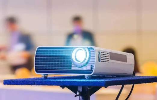 White projector on a blue stand projecting light in a blurred indoor setting with people.