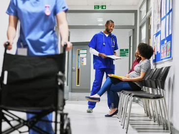 Healthcare worker in blue scrubs talks to seated patient in hospital corridor with another staff passing by.