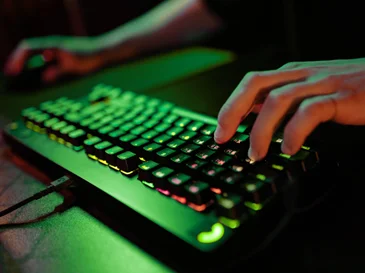 Close-up of hands typing on a backlit gaming keyboard with RGB lighting on a dark desk.