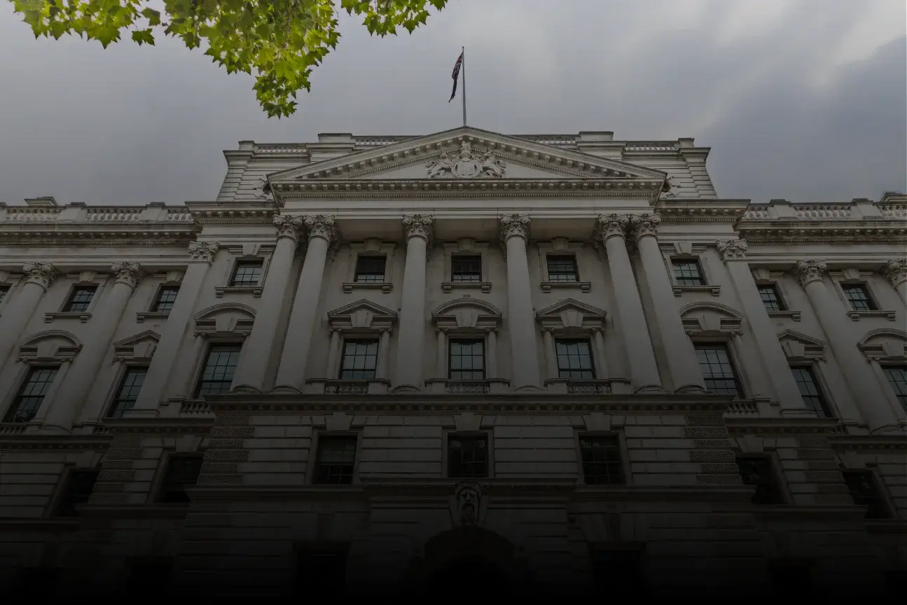 Front facade of a classical government building with columns and a flag on top under gray sky.
