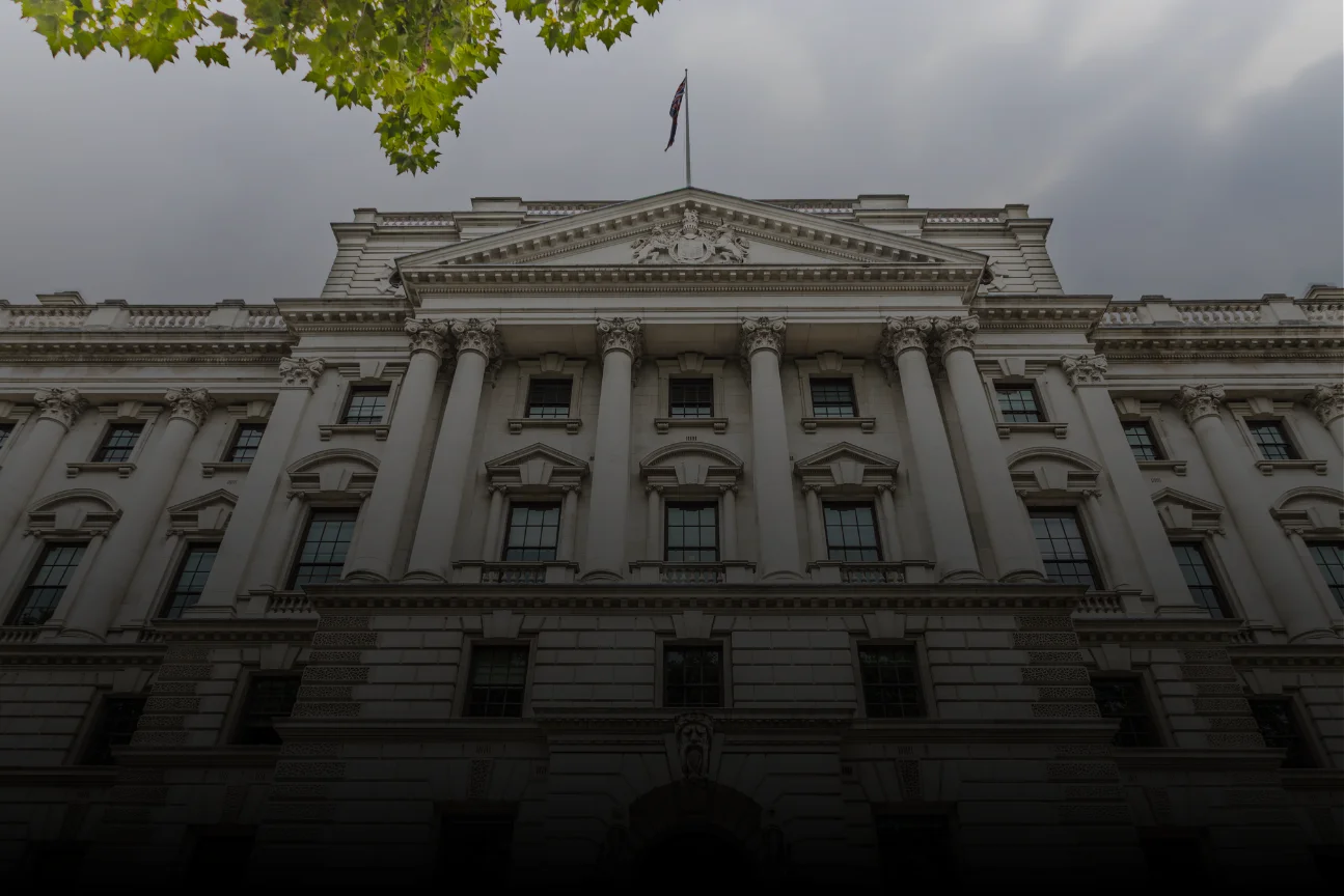 Front facade of a classical government building with columns and a flag on top under gray sky.