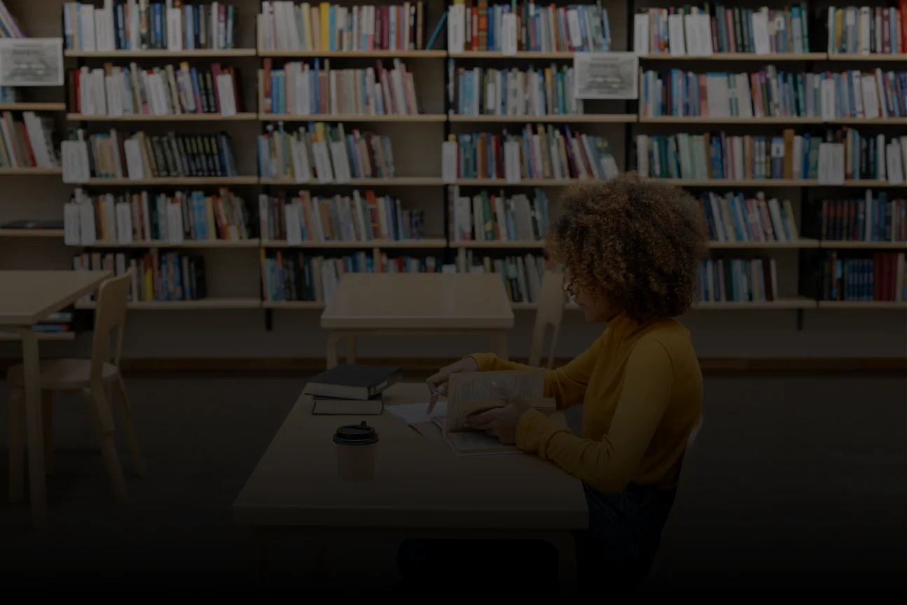Person reading a book at a library table with bookshelves in the background and a coffee cup nearby.