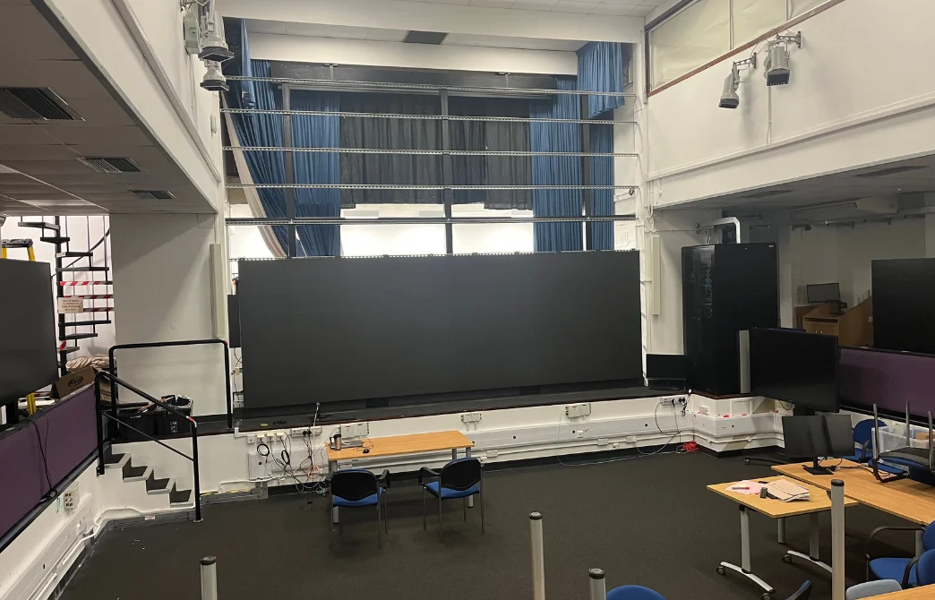 Large indoor room with a big black LED screen, desks, chairs, and equipment around it.