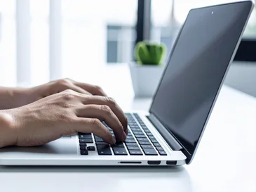 Person typing on a laptop keyboard near a window with a small potted plant in the background.