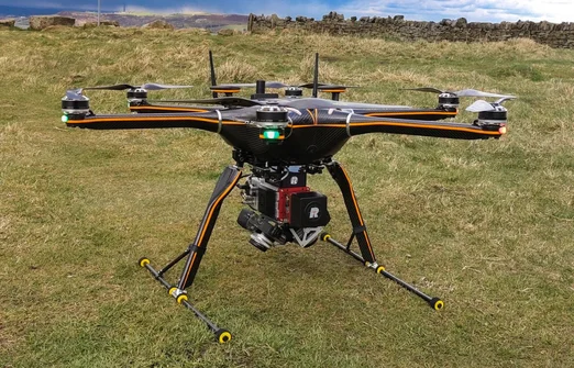 Black and orange drone with six propellers and a camera, flying over grassy field near stone wall.